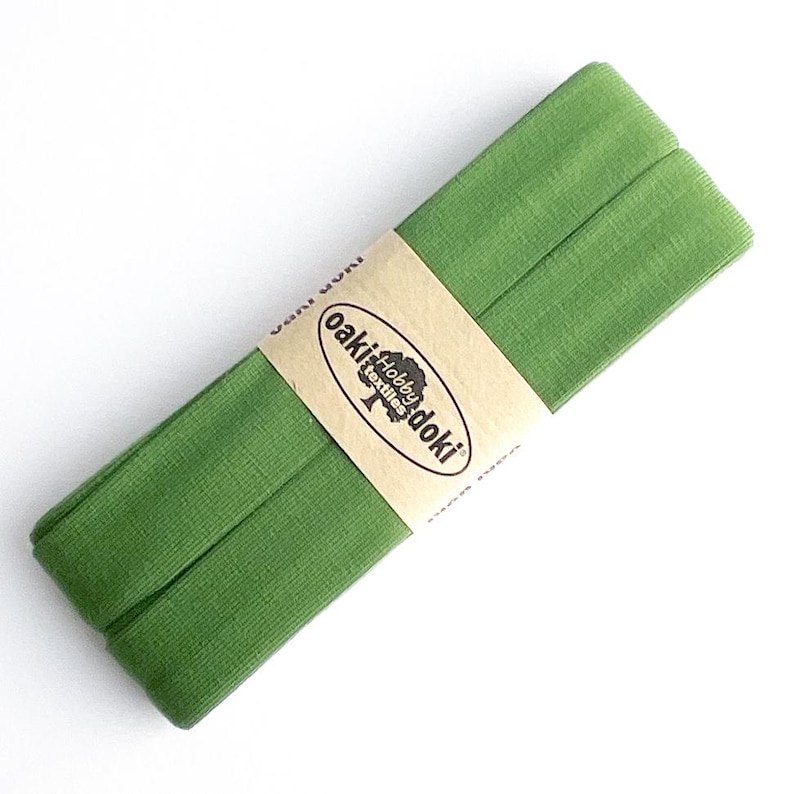 May include: A folded length of green fabric ribbon secured with a tan paper band. The band features the black printed text "oaki doki hobby textiles" along with a small tree icon. The fabric has a fine, textured, and slightly sheer appearance.