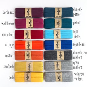 May include: Fourteen rolls of folded fabric binding tape arranged in two columns. Each roll is secured with a tan paper band featuring the Oaki Doki logo. The colors include bordeaux, berry, red, orange, rust, mustard, yellow, dark petrol, petrol, turquoise, royal blue, dark gray heather, gray heather, and light gray heather.
