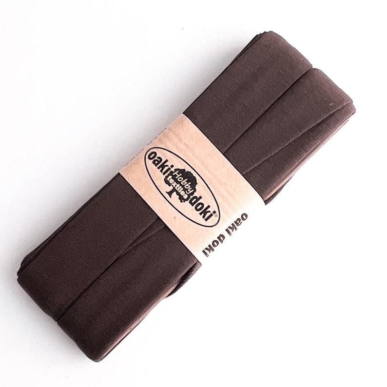 May include: A folded roll of dark brown bias tape secured with a light tan paper band. The band features the text oaki doki hobby textiles and the brand name oaki doki printed in black ink. The fabric has a smooth, matte, and uniform texture.