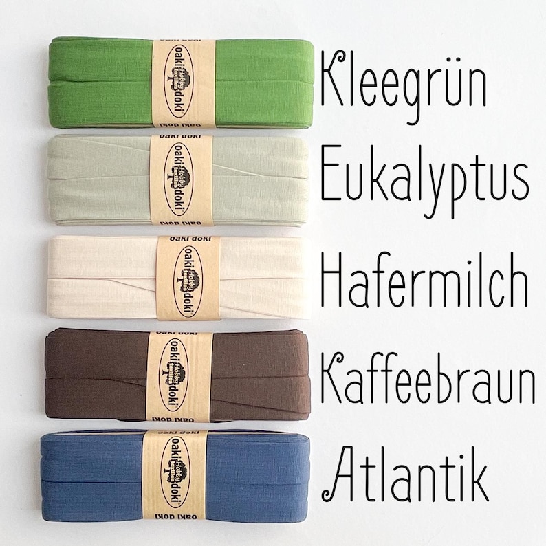 May include: Five folded fabric strips in clover green, eucalyptus, oat milk, coffee brown, and atlantic blue. Each strip is secured with a tan paper band featuring the text oaki doki and a tree logo. The corresponding color names are printed in black text.