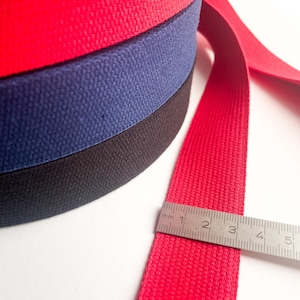 May include: Three rolls of woven cotton webbing in red, navy blue and black. The red webbing is being measured with a ruler, showing 5 centimetres.
