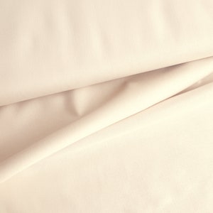 Könnte beinhalten: Nahaufnahme eines glatten, cremefarbenen Stoffes mit einer dezenten Textur. Der Stoff ist so gefaltet, dass er weiche, fließende Linien erzeugt.