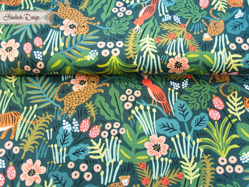 May include: A dark green fabric featuring a repeating pattern of orange leopards, tigers, and birds amidst pink flowers, white berries, and various green tropical leaves. A small rectangular label with the text H&auml;ndisch-Design is visible.