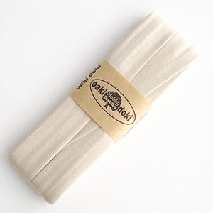 May include: A folded strip of light beige fabric bias tape secured with a tan paper band. The band features the text oaki doki and a logo with the words Hobby textiles. The fabric has a visible woven texture and is arranged in a long, flat stack.