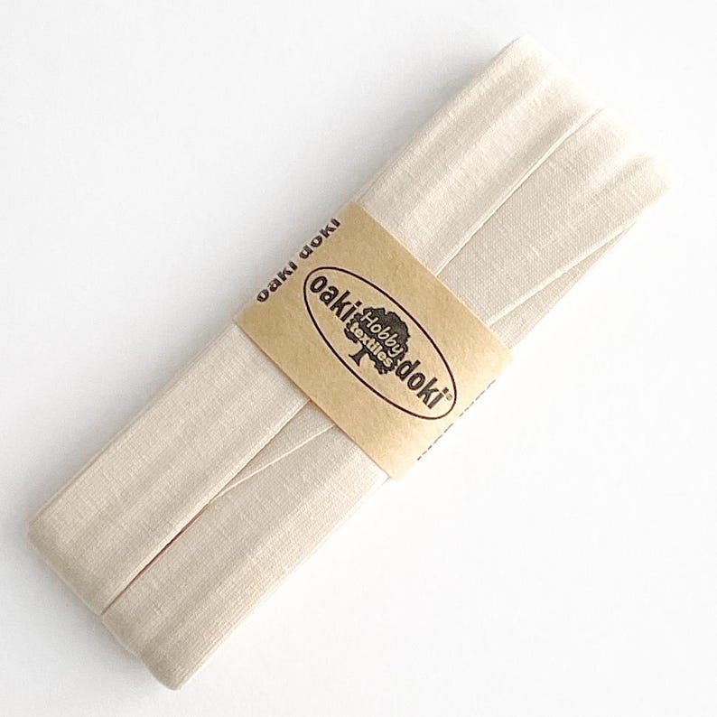 May include: A folded strip of light beige fabric bias tape secured with a tan paper band. The band features the text oaki doki and a logo with the words Hobby textiles. The fabric has a visible woven texture and is arranged in a long, flat stack.