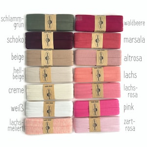 May include: Fourteen rectangular rolls of folded fabric elastic arranged in two columns. Each roll is secured with a tan paper band featuring the text oaki doki and a tree logo. The colors include shades of green, brown, beige, cream, white, and various pinks.