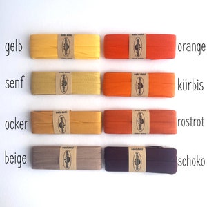 May include: Eight rectangular spools of folded fabric bias tape arranged in two columns. The colors include yellow, mustard, ochre, beige, orange, pumpkin, rust red, and dark chocolate brown. Each spool features a tan paper label with the text oaki doki.