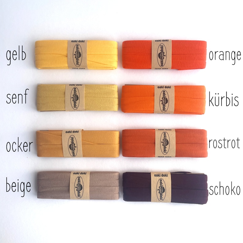 May include: Eight rectangular spools of folded fabric bias tape arranged in two columns. The colors include yellow, mustard, ochre, beige, orange, pumpkin, rust red, and dark chocolate brown. Each spool features a tan paper label with the text oaki doki.