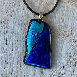 Peut inclure: Un collier pendentif en verre dichroïque avec un cordon noir. Le pendentif est de forme rectangulaire avec un bord noir. Le verre présente des nuances de bleu et de turquoise, avec un motif texturé. Le pendentif est fixé à une bélière argentée.