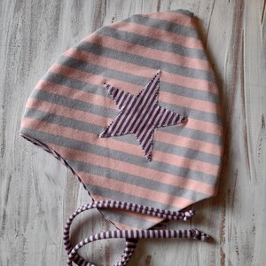 May include: A pink and gray striped baby bonnet with a star applique. The star is made of a darker pink striped fabric. The bonnet has matching striped ties.