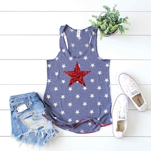 Puede incluir: Un top azul de tirantes con una estrella roja de lentejuelas sobre un patr&oacute;n de estrellas blancas. El top est&aacute; sobre una superficie de madera blanca con un par de zapatillas blancas y una planta en maceta.