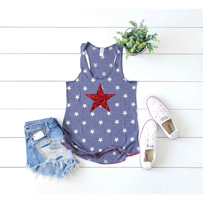 Puede incluir: Un top azul de tirantes con una estrella roja de lentejuelas sobre un patr&oacute;n de estrellas blancas. El top est&aacute; sobre una superficie de madera blanca con un par de zapatillas blancas y una planta en maceta.