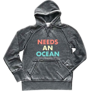 May include: Dark gray hooded sweatshirt with the text "NEEDS AN OCEAN" in red, yellow, and teal. The sweatshirt has a front pocket and a drawstring hood. The fabric has a distressed, vintage look.