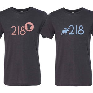 May include: Two dark gray t-shirts. One shirt has the number "218" in pink with a pink circle and a silhouette of the state of Minnesota. The other shirt has the number "218" in light blue with a light blue moose silhouette.