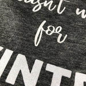 May include: Close-up of a gray shirt with white glitter lettering that reads "wasn't made for winter".
