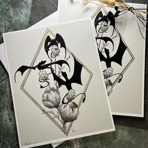 May include: Two black and white prints of a stylised dragon with floral elements. The dragon is depicted in a diamond shape with a gold outline. The prints are on white paper with a textured surface.