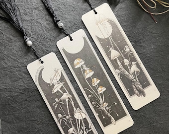 May include: Three illustrated bookmarks with black tassels. Each bookmark features a different mushroom design in shades of gray, white, and tan. The bookmarks have a crescent moon and star design at the top. The tassels are black with a decorative bead.