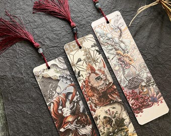 May include: Three bookmarks with intricate illustrations of mythical creatures. Each bookmark features a different creature, including a fox, an owl, and a dragon. The bookmarks are made of thick paper and have a cream-colored background. Each bookmark has a red tassel attached to it.