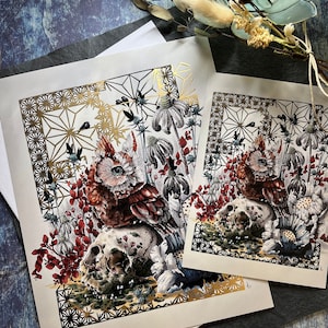 May include: A detailed illustration of an owl perched on a skull, surrounded by flowers and foliage. The artwork is framed with a gold geometric pattern.