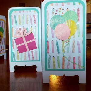 May include: A multi-panel birthday card with a teal border. The card features a pink gift box with a white bow, and a cluster of pastel balloons with a white string.