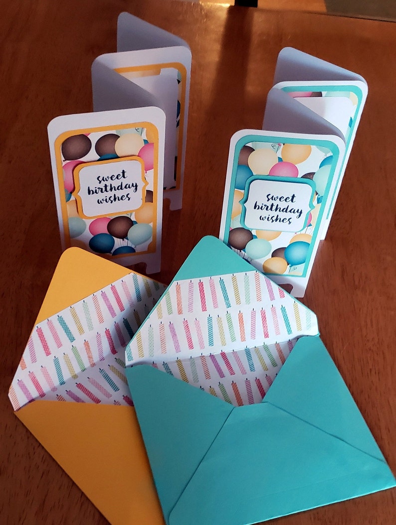 May include: Two folded birthday cards with a balloon and "sweet birthday wishes" design. The cards are sitting on top of two envelopes with a colorful candle pattern.
