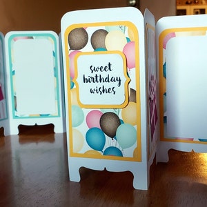 May include: A white card with a yellow border and a colorful balloon pattern. The card is folded into a screen shape with four panels. The center panel has a white banner with the text "Sweet birthday wishes".