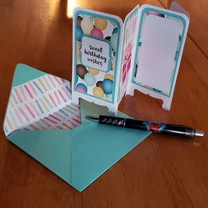 May include: A teal blue card with a pop-up design featuring balloons and the text "sweet birthday wishes". The card is open and folded to reveal a blank white space on the inside. A teal blue envelope and a black pen are also visible.