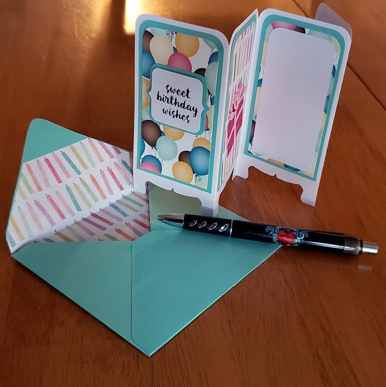May include: A teal blue card with a pop-up design featuring balloons and the text "sweet birthday wishes". The card is open and folded to reveal a blank white space on the inside. A teal blue envelope and a black pen are also visible.