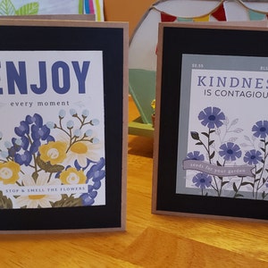 May include: Two framed cards with floral designs and inspirational messages. The left card has a yellow and blue floral design with the text "ENJOY every moment STOP & SMELL THE FLOWERS". The right card has a blue floral design with the text "50.55 BLUE VARIETY KINDNESS IS CONTAGIOUS seeds for your garden".
