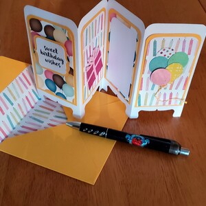 May include: A white card with a yellow border and a colorful birthday theme. The card is folded into four panels. The front panel has a colorful balloon design and the text "sweet birthday wishes". The second panel has a colorful gift box design. The third panel is blank. The fourth panel has a colorful candle design with balloons. The card is sitting on a yellow envelope with a colorful candle design.