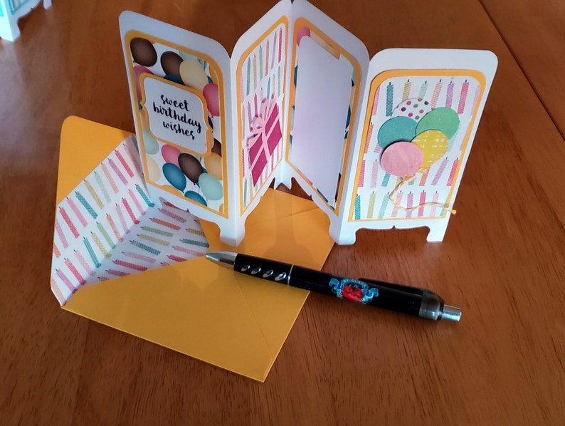 May include: A white card with a yellow border and a colorful birthday theme. The card is folded into four panels. The front panel has a colorful balloon design and the text "sweet birthday wishes". The second panel has a colorful gift box design. The third panel is blank. The fourth panel has a colorful candle design with balloons. The card is sitting on a yellow envelope with a colorful candle design.