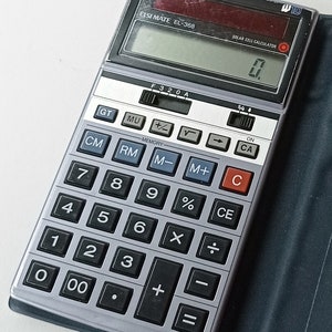 Könnte beinhalten: Ein silberner und schwarzer Sharp ELSI MATE EL-368 Solarzellen-Taschenrechner. Der Rechner hat ein Display mit "0" und zahlreiche Tasten für mathematische Funktionen. Er befindet sich in einem dunkelblauen Etui.