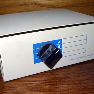 Könnte beinhalten: Ein rechteckiger, hellgrauer Datenübertragungsschalter mit einem schwarzen Knopf und blauen Etiketten. Der Schalter hat die Beschriftungen A und B, und der Text "DATA TRANSFER SWITCH" ist aufgedruckt.
