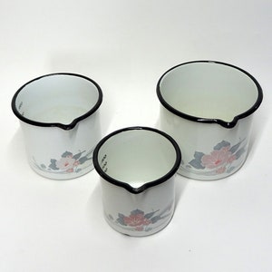 Könnte beinhalten: Set aus drei weißen Emaille-Messbechern mit schwarzen Rändern und Ausgüssen. Jeder Becher hat ein Blumendesign mit rosa und grauen Blüten. Die Becher haben Messmarkierungen in Millilitern.