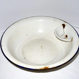 Könnte beinhalten: Ein weißes Emaille-Waschbecken mit blauem Rand und einer kleinen, angebrachten Seifenschale. Das Waschbecken weist Alterserscheinungen mit Rostflecken und Abnutzung auf. Das Design ist schlicht und funktional, typisch für Vintage-Geschirr.
