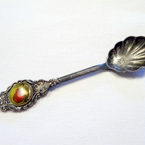 Könnte beinhalten: Ein silberner Vintage-Löffel mit einer muschelförmigen Schale und einem verzierten Griff. Der Griff hat ein kleines, rundes Bild mit einem roten Fruchtdesign. Der Löffel ist wahrscheinlich ein Souvenir oder ein Sammlerstück.