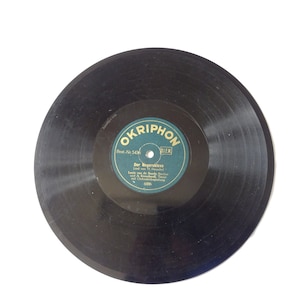 May include: A vintage black vinyl record with a teal and white label. The label reads "OKRIPHON" and features German text. The record is a classic music format, likely for a record player.