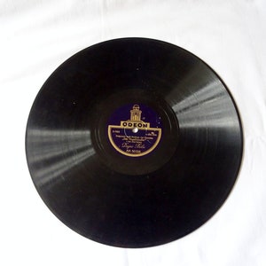 May include: A vintage black vinyl record with a purple and gold label. The label features the word "ODEON" and text in a foreign language. The record is set against a white background.