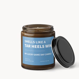 Op de afbeelding: Een bruine glazen potkaars met een zwart deksel, met het opschrift "Smells Like a Tar Heels Win". De kaars heeft een blauw etiket met witte tekst, en de tekst "My Lucky Game Day Candle". De kaars is 312 gram.