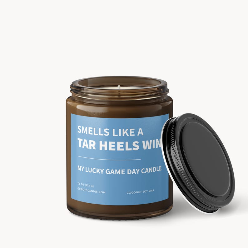 Op de afbeelding: Een bruine glazen potkaars met een zwart deksel, met het opschrift "Smells Like a Tar Heels Win". De kaars heeft een blauw etiket met witte tekst, en de tekst "My Lucky Game Day Candle". De kaars is 312 gram.