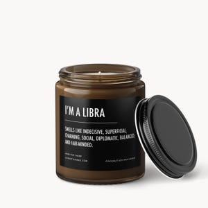May include: A brown glass jar candle with a black label and lid. The label reads "I'M A LIBRA" with a description of the scent. The lid is off to the side. The candle is a coconut soy wax candle.