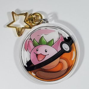 May include: A clear acrylic keyring featuring a cartoon character inside a stylized ball. The character is pink with green leaves and a smiling face. The keyring has a gold star-shaped clasp.