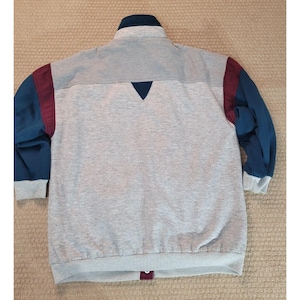 May include: A gray zip-up jacket with a navy blue collar and sleeves, and burgundy accents. The jacket has a ribbed texture on the upper back and a triangular navy blue design element. The cuffs and hem are gray.