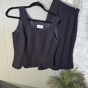 May include: A two-piece dark navy suit set, including a sleeveless top and a skirt, hanging on a black hanger. The top has a scoop neckline and a unique scalloped hemline. The skirt is a straight, knee-length style. The label reads "ALBERT NIPON".