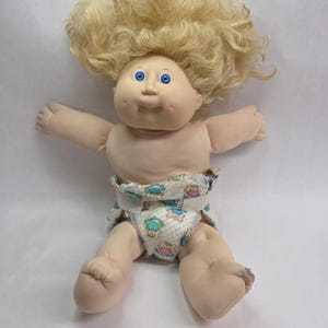 May include: A vintage baby doll with blonde, curly hair and blue eyes. The doll is wearing a diaper with a pattern of teddy bears and other designs. The doll's arms and legs are outstretched.