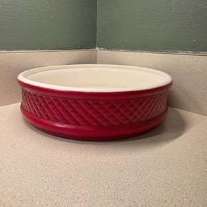 May include: A red ceramic oval baking dish with a white interior. The exterior features a diamond pattern. The dish is sitting on a light-colored countertop. The dish is approximately 12 inches in diameter.