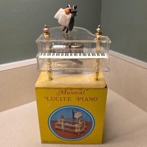 May include: A clear lucite miniature grand piano music box with gold-toned legs and a keyboard, topped with a dancing couple figurine. It sits on a yellow cardboard box labeled Musical LUCITE PIANO with a circular image of the piano.