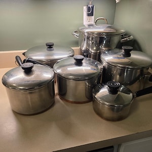 May include: A collection of five stainless steel cooking pots and pans of varying sizes, each featuring a polished metallic finish, a matching lid with a black knob, and a black handle. The set includes a large stockpot and four smaller saucepans.
