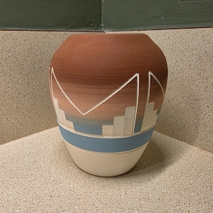 May include: A rounded ceramic vase featuring a gradient color scheme that transitions from a deep terracotta brown at the top to a soft cream at the base. The surface is decorated with carved geometric patterns and a horizontal blue band.