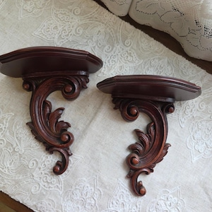 May include: Two dark brown wooden wall shelves featuring a semi-circular top surface and an ornate, carved bracket support with scrolling leaf and vine motifs. The shelves have a smooth, polished finish and are designed for mounting on a flat wall.